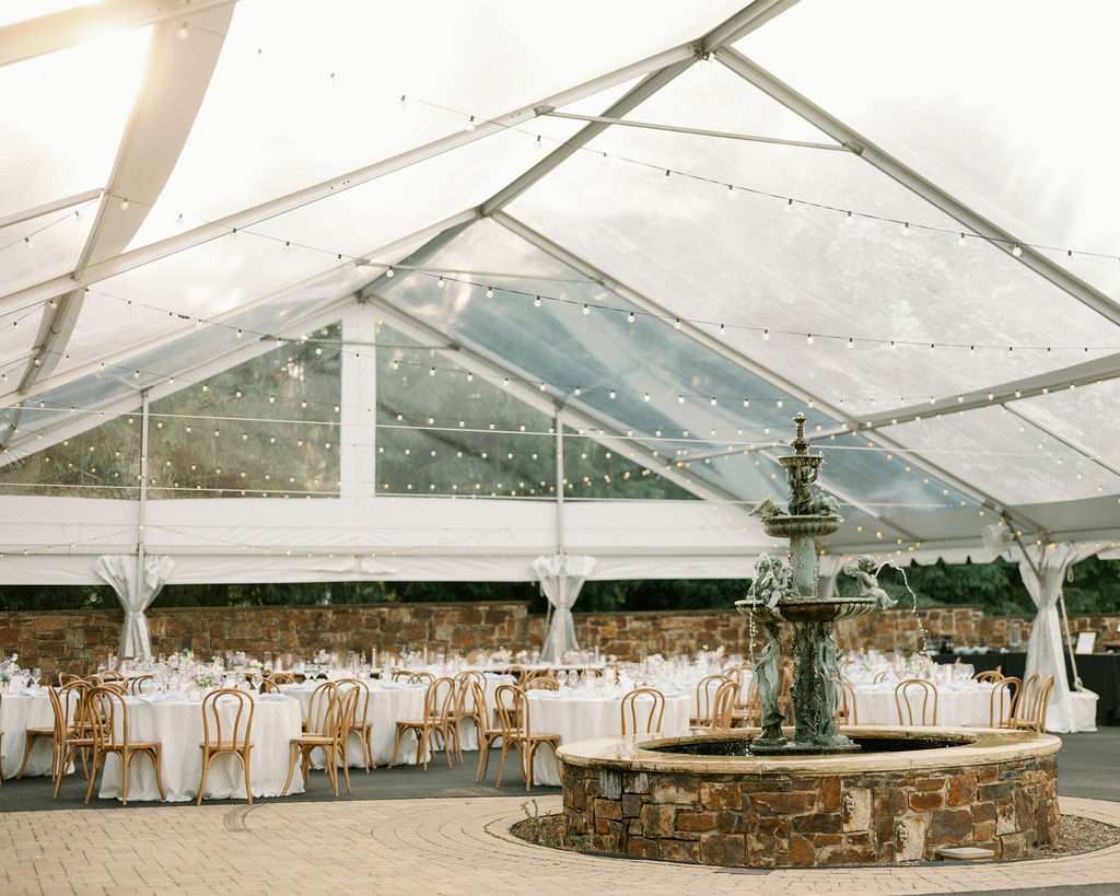 Clear tented wedding reception with large fountain in the middle and white tables with brown chairs